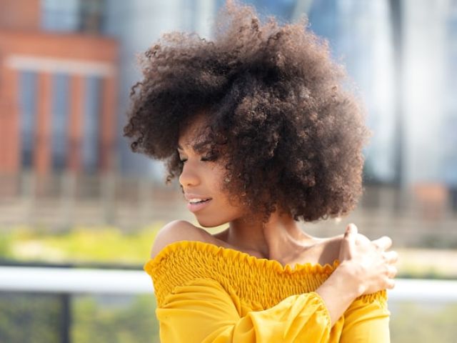 How Creatine Became an Anti-Aging and Longevity Essential 9 Young woman with voluminous natural curly hair in bright yellow off-shoulder top poses outdoors against blurred urban backdrop.